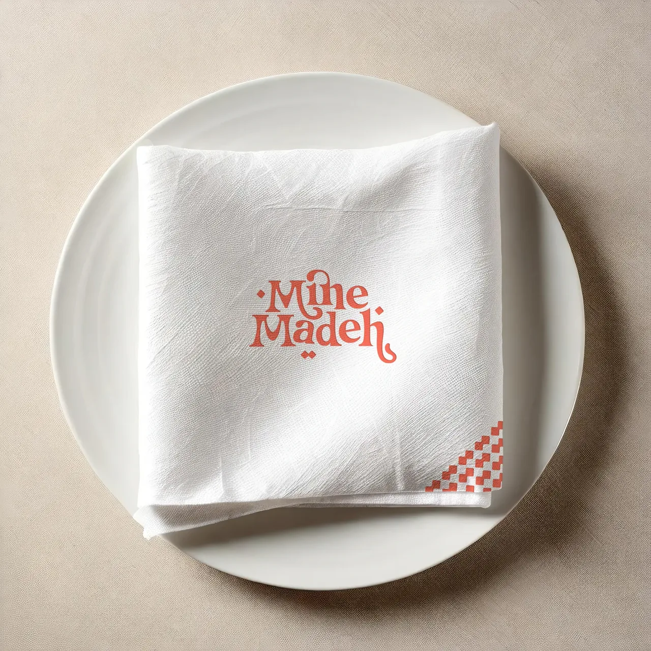 Serviette blanche pliée sur une assiette, logo Mine Madeh corail et motif de losanges en coin